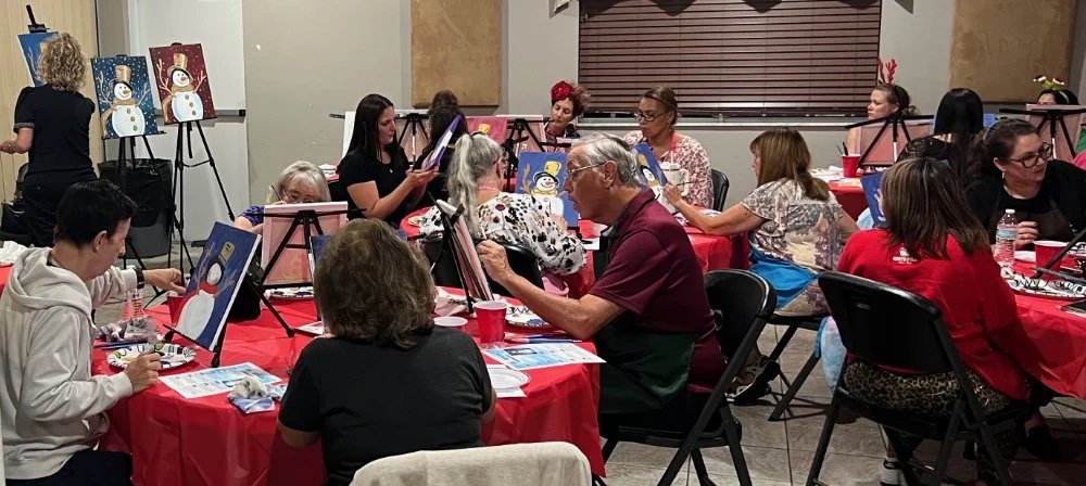 People participating in a holiday-themed painting class, painting snowmen on canvases at tables covered with red tablecloths.