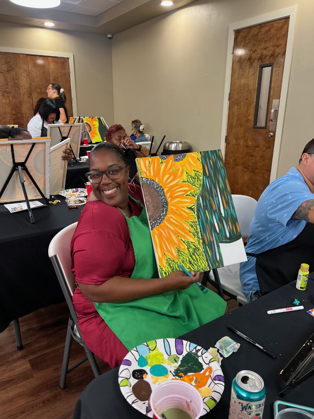 Woman smiling and holding a sunflower painting at an art event, seated at a table with paint supplies, in a room with other people painting.