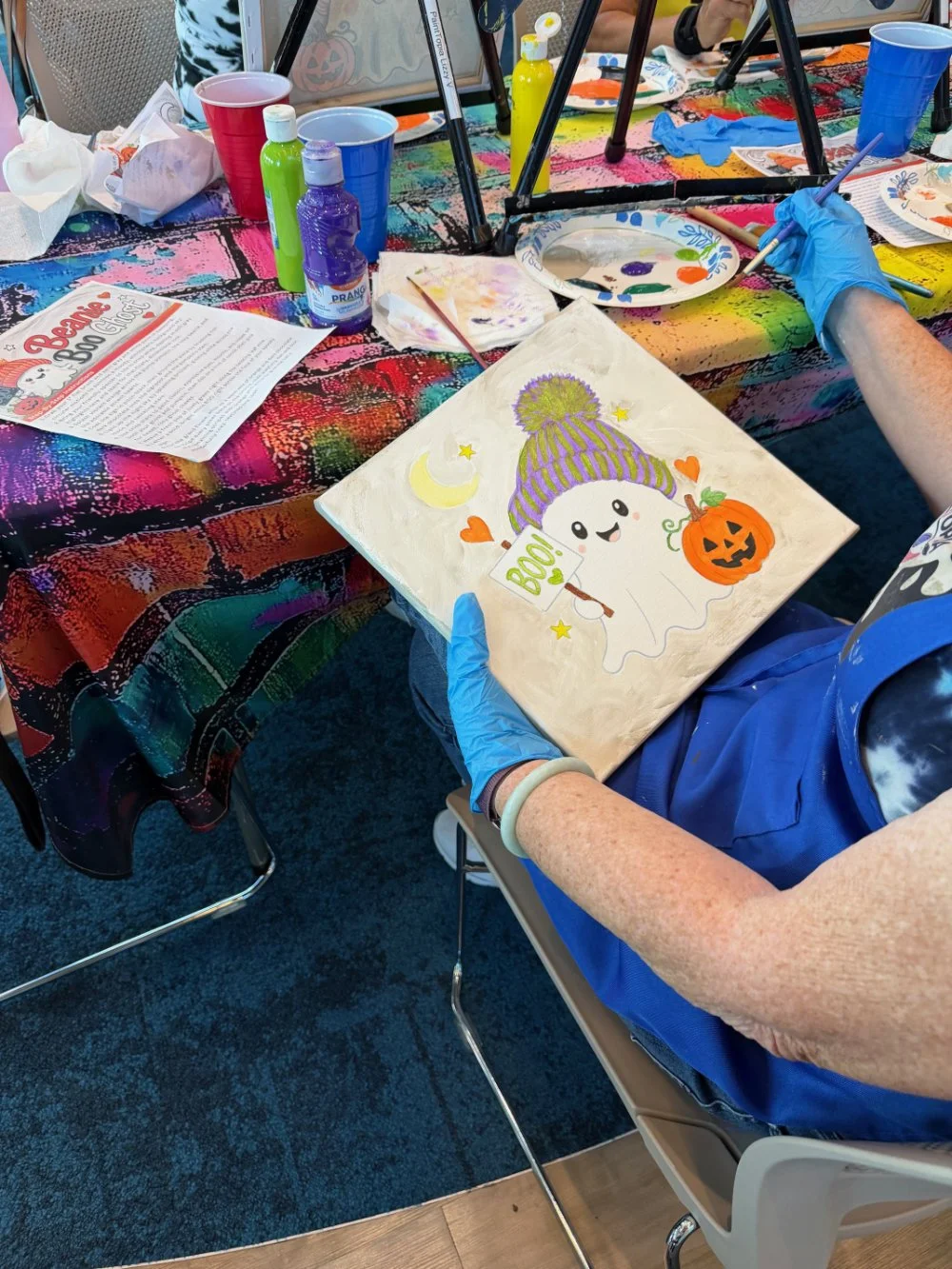 A person wearing blue gloves painting a Halloween-themed picture featuring a cute ghost with a purple and yellow hat, holding a pumpkin-shaped bucket, surrounded by stars, hearts, a crescent moon, and a small sign reading "Boo!" on a colorful, splatt