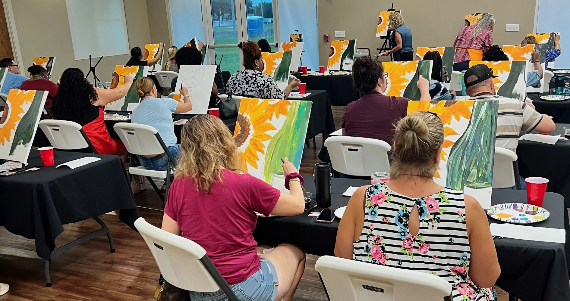 People participating in a group painting class, creating sunflower paintings on canvases in a well-lit room with large windows.