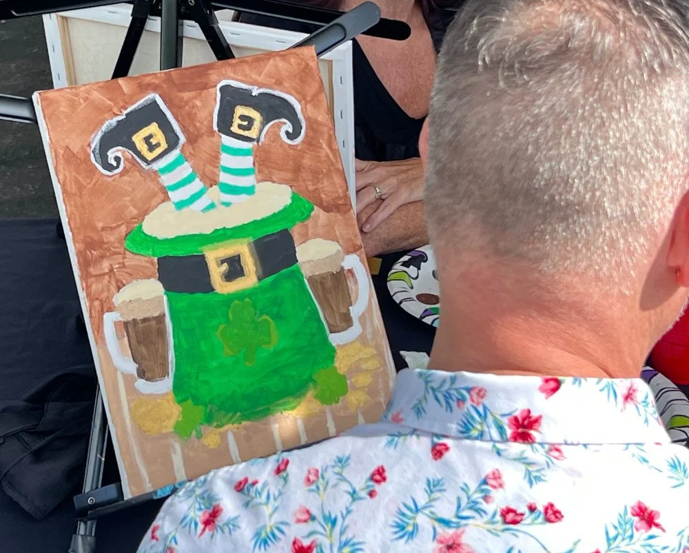 A person with short gray hair, wearing a white shirt with a red floral pattern, looks at a colorful painting of a leprechaun with striped socks and a pot of gold, likely for a St. Patrick's Day celebration.