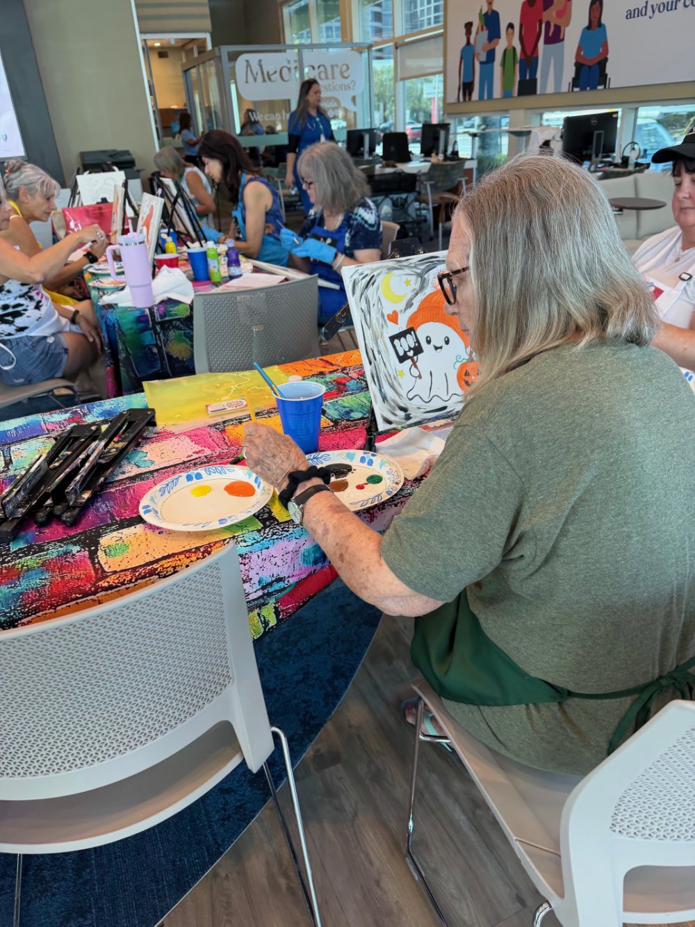 People painting at a table during a group art activity, with an artist painting a Halloween-themed picture of a ghost, pumpkin, and moon.