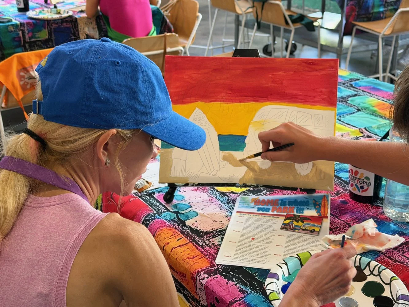 Two women are painting a landscape on a canvas at an indoor art event. One woman is wearing a blue cap and a pink sleeveless shirt, and the other is using a paintbrush. The table has a colorful tablecloth, paint supplies, papers, and a coffee cup.