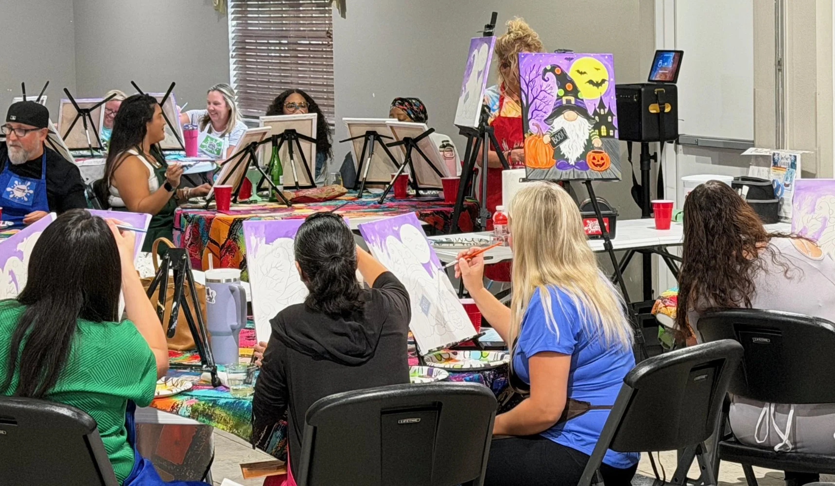 People participating in a group painting class, with canvases depicting Halloween-themed scenes, in a room with tables, chairs, and art supplies.