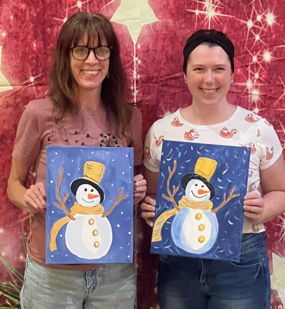 Two women smiling and holding paintings of snowmen in front of a festive red background with white star-like designs.