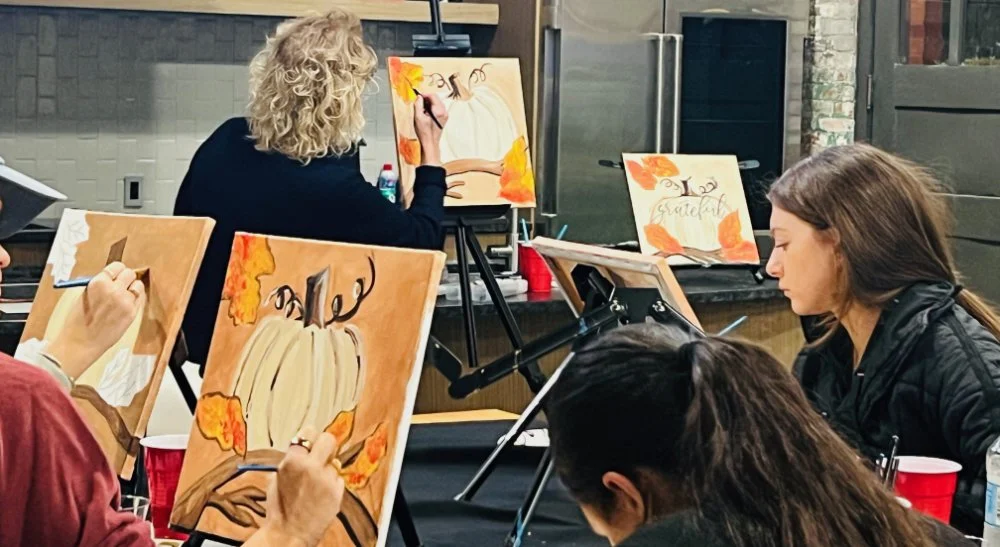 People painting autumn-themed canvases featuring pumpkins and orange leaves in a group art class.
