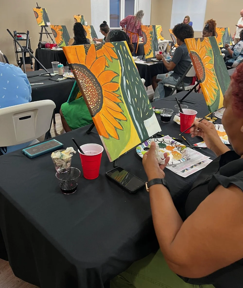People participating in a painting class, creating sunflower paintings on canvases set up on tables covered with black tablecloths, with art supplies and drinks on the tables.