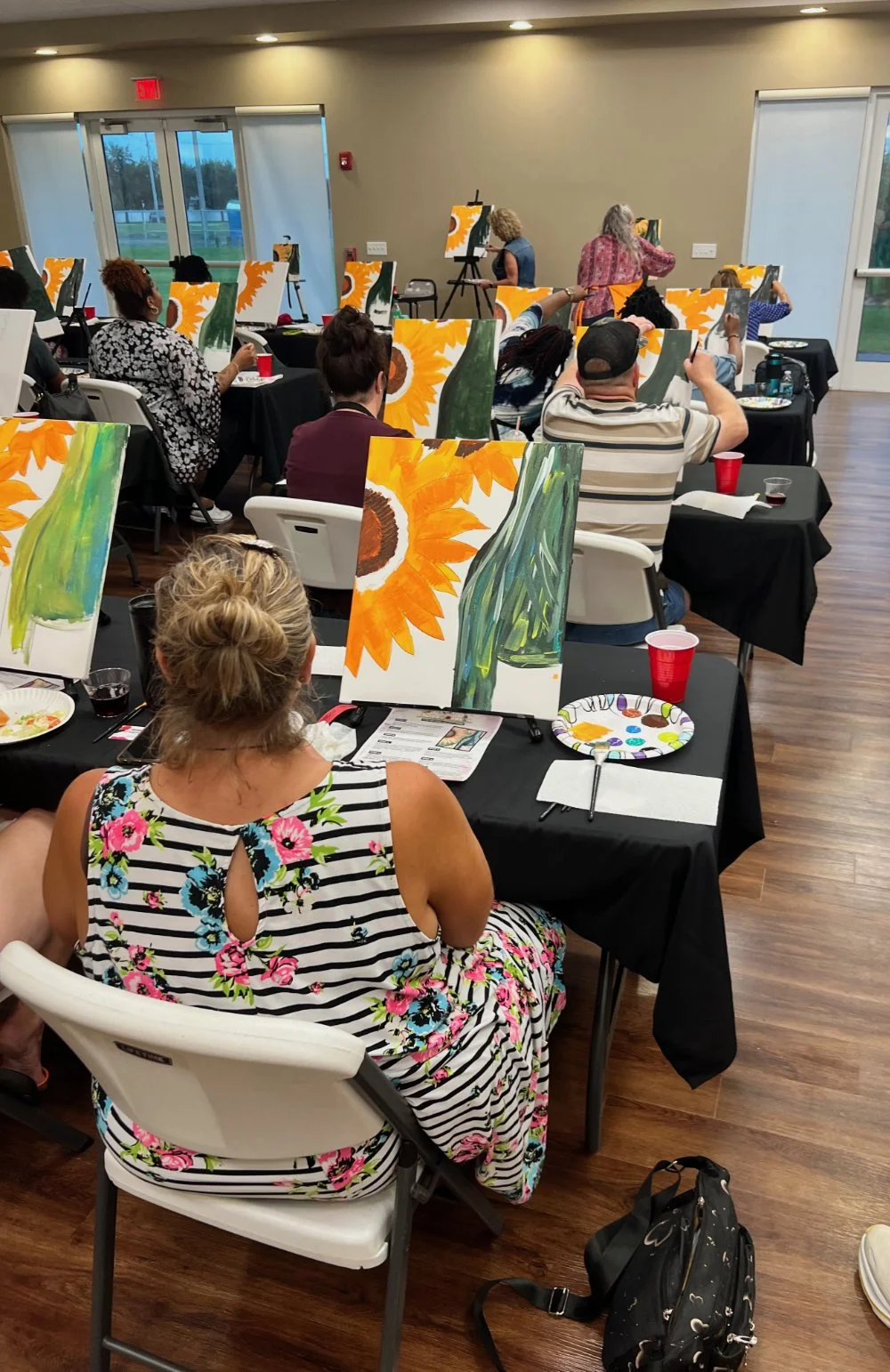 People participating in a group painting class, sitting at tables with canvases and painting supplies, creating sunflower paintings in a well-lit room with large windows.