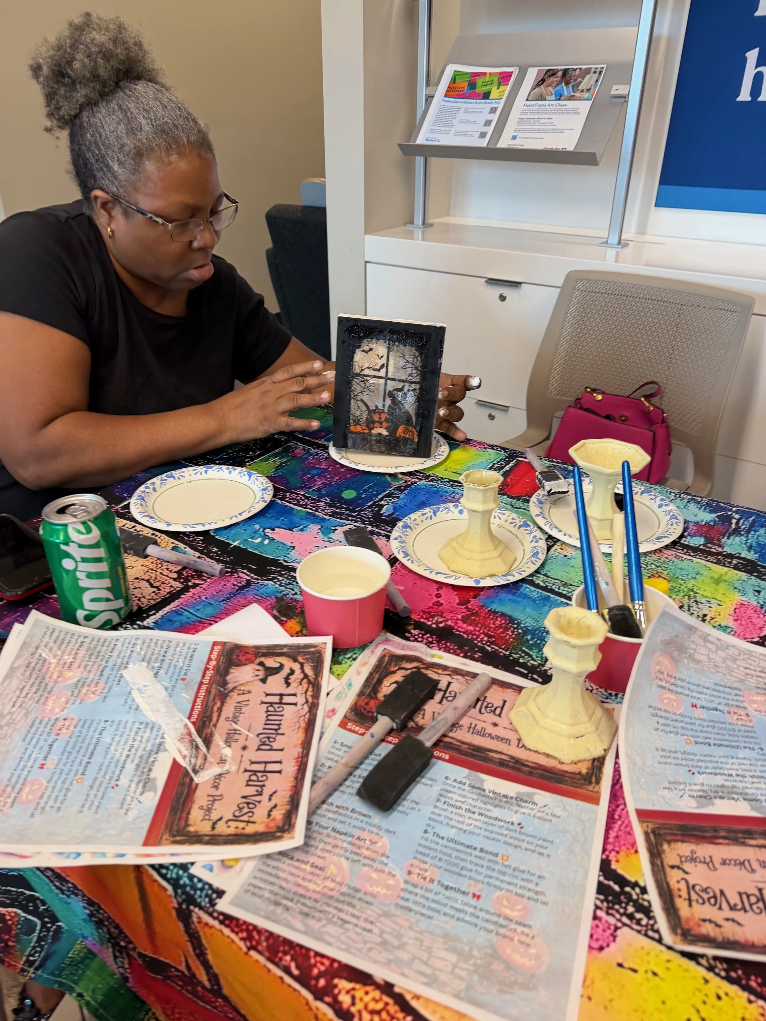 A woman with glasses and gray hair styled in a bun is sitting at a colorful table working on a Halloween-themed craft project. The table is covered with newspapers, paint supplies, and Halloween decorations, including painted pumpkins and various art
