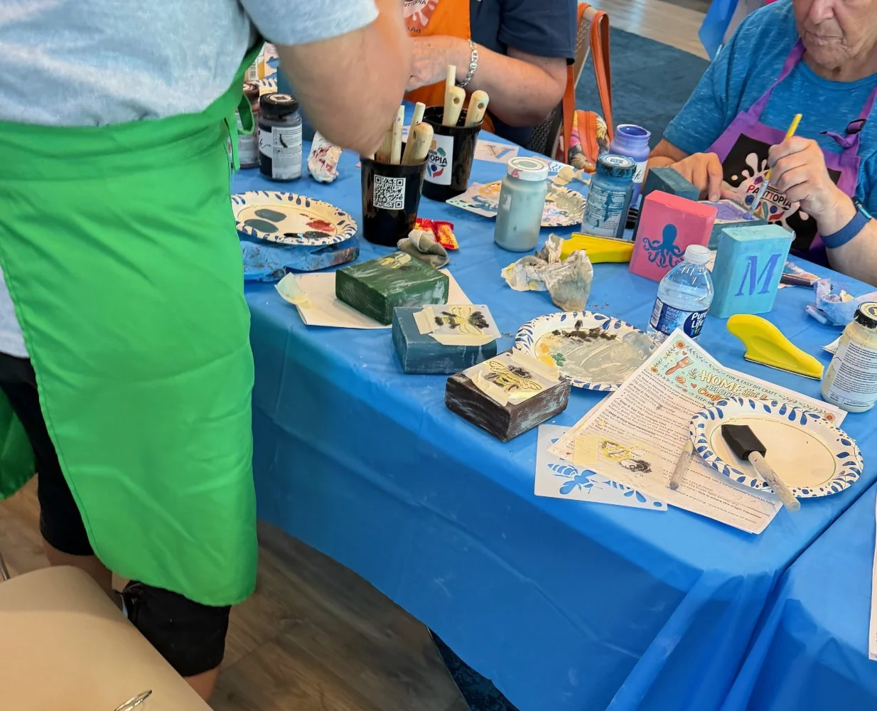People engaging in a crafting activity, painting and decorating objects such as blocks and plaques, with art supplies on a table covered with a blue tablecloth.