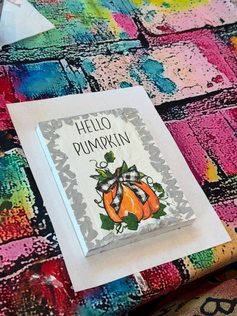 A decorative sign with a picture of a pumpkin, leaves, and a black-and-white checkered bow. The sign says 'Hello Pumpkin' and is placed on a colorful, multicolored textured surface.