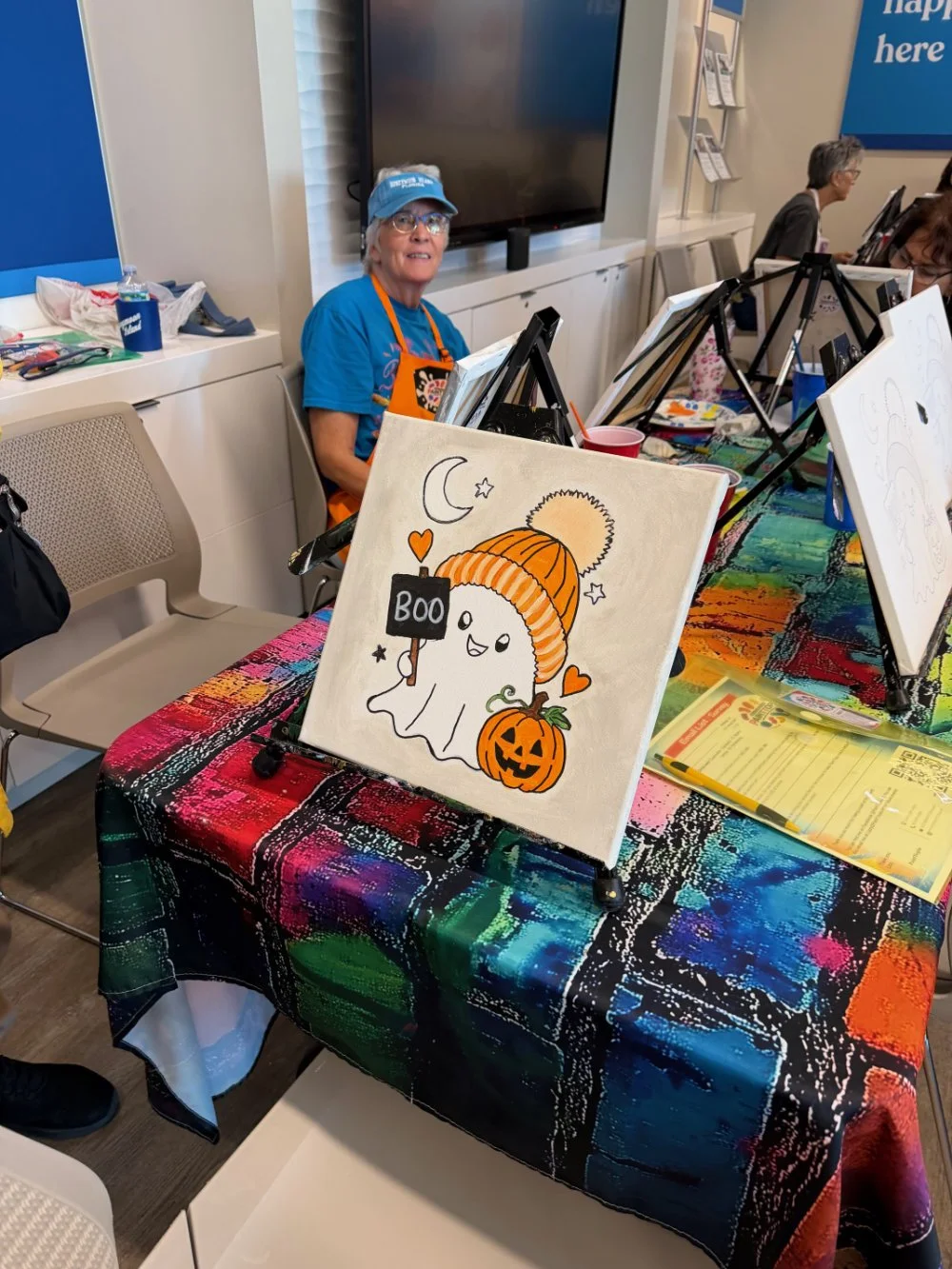 A woman in a blue cap and shirt sitting behind a table, which displays Halloween-themed artwork of a ghost holding a sign that says 'BOO,' a pumpkin, a moon, and stars, with colorful tablecloths and art supplies around her.