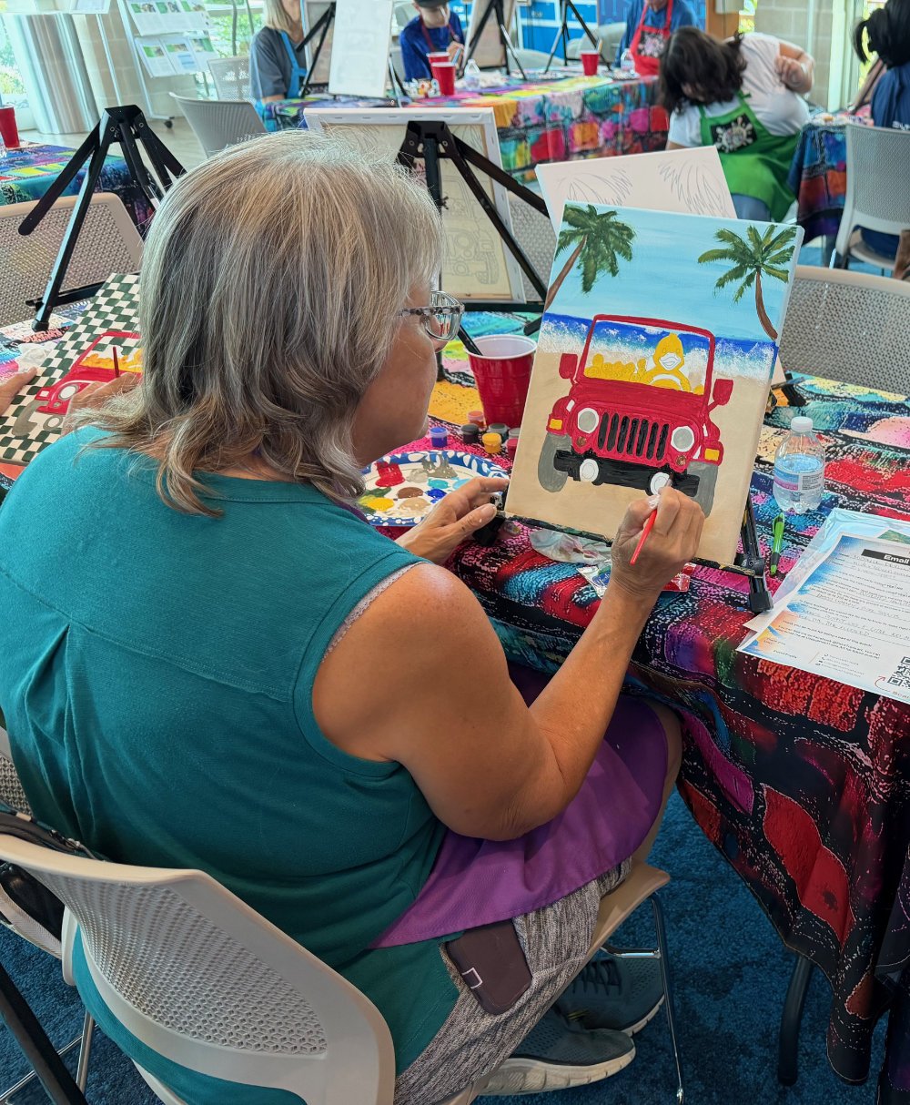 A woman with gray hair and glasses painting a picture of a red Jeep at an outdoor-themed art class, with palm trees, a sandy beach, and a blue sky in the background.