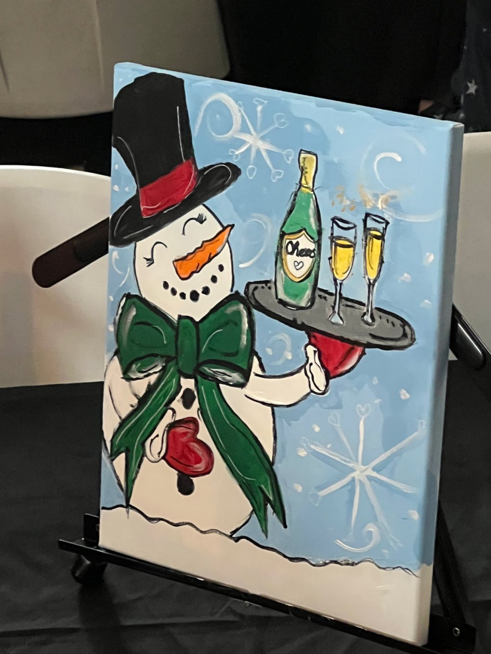A snowman with a black top hat with a red band, orange carrot nose, and a large green bow tie. The snowman is holding a tray with a bottle of champagne and two glasses. The background is light blue with snowflakes.