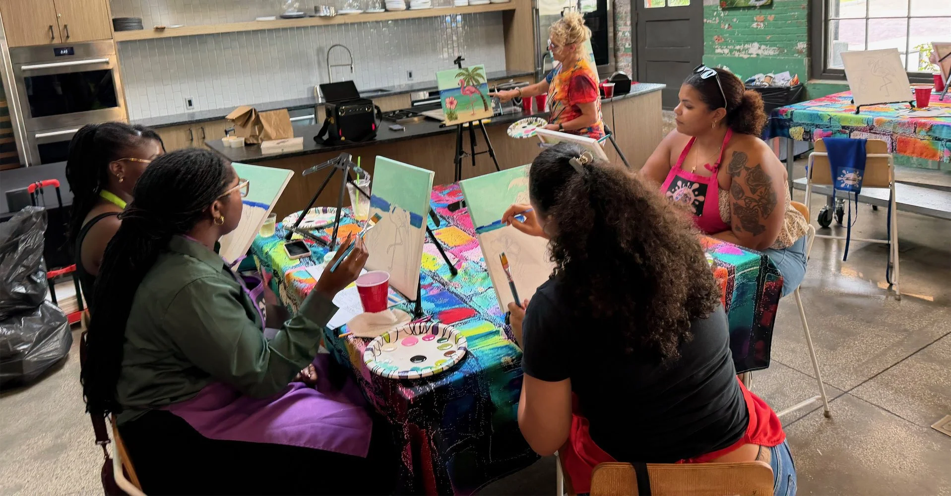 A group of women painting on canvases seated around a table covered with a colorful tablecloth in an art studio.