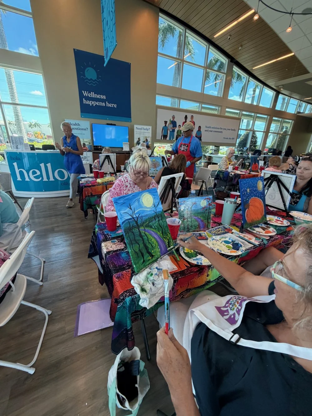 Participants, mostly elderly women, engaged in a painting activity at a wellness center with colorful paintings on easels, some with nature themes like a rainbow and a moon, in a bright room with large windows and a high ceiling.