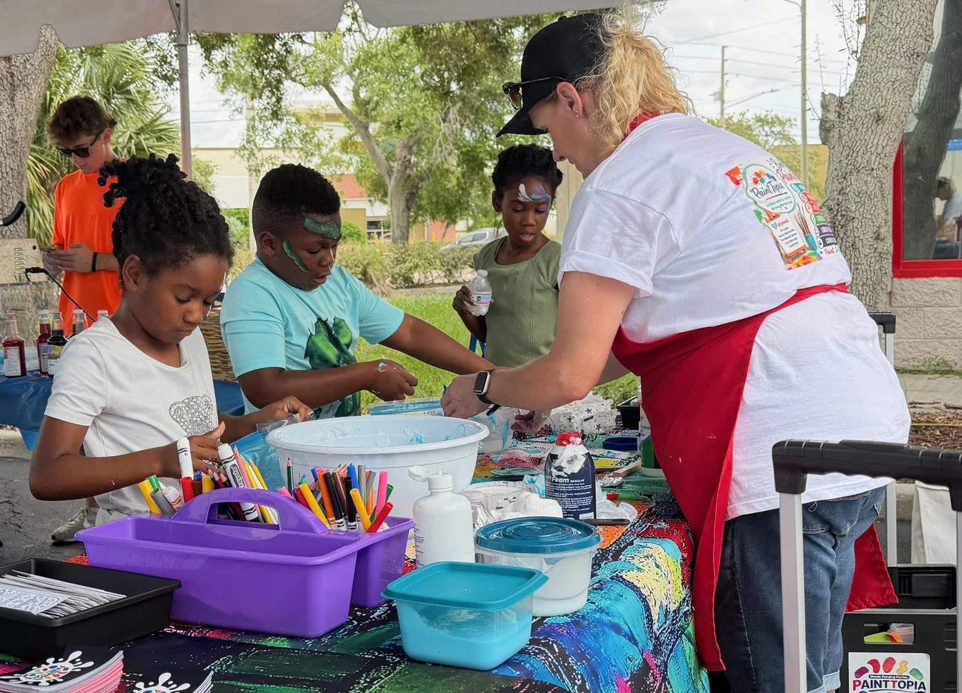 Children participating in face painting activity at an outdoor event, with a woman assisting at a table filled with art supplies and face paints.