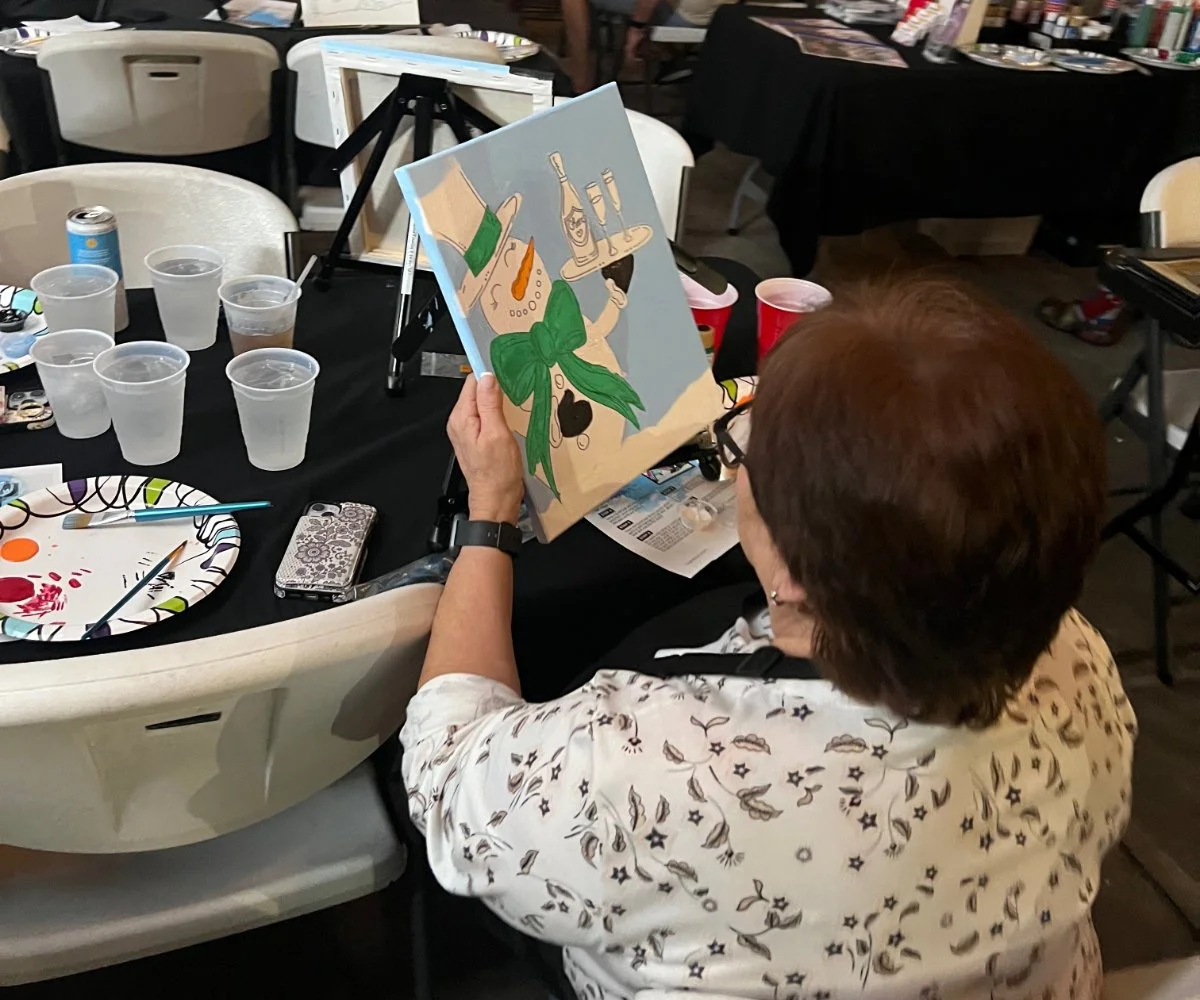 A woman with short brown hair and glasses is sitting at a table, painting a colorful artwork of a snowman with a green scarf. The table has multiple cups of water, a paper plate with paint, a paintbrush, a phone, and a red cup. In the background, the