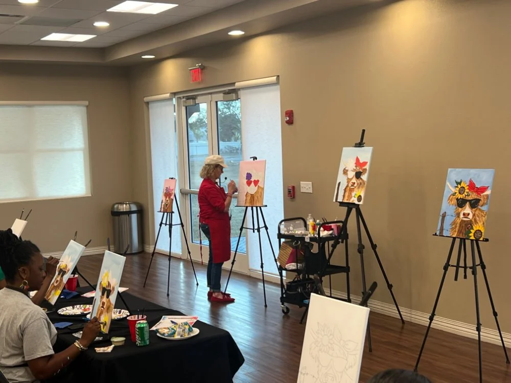 People participating in a painting class, working on paintings of a dog with sunglasses and flowers, in a room with beige walls, windows, and a wooden floor.