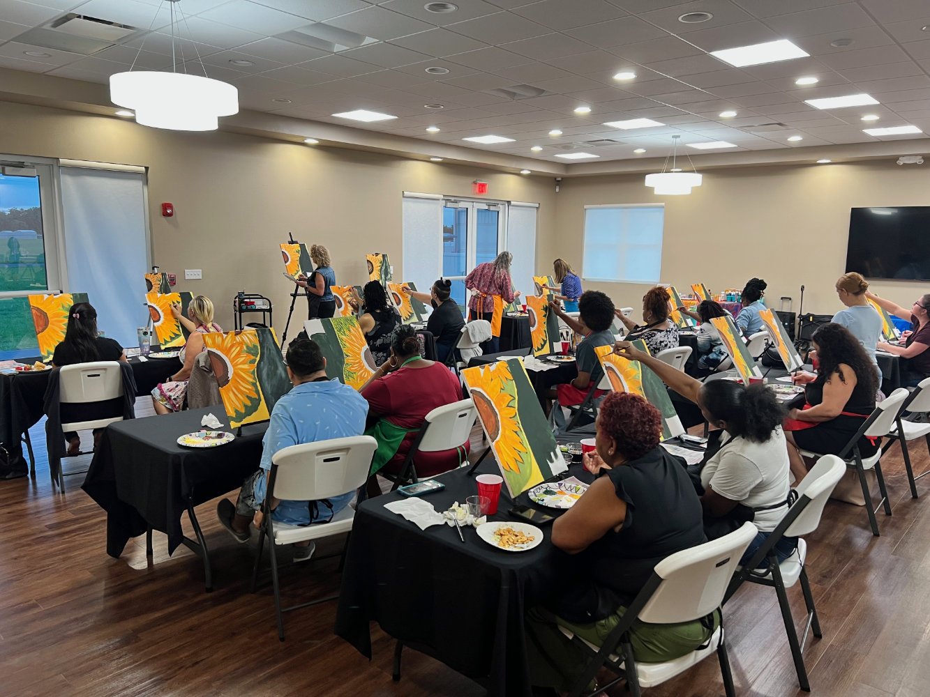 A group of people participating in a painting class, sitting at tables with canvases of sunflowers, in a well-lit room with windows and a large TV on the wall.