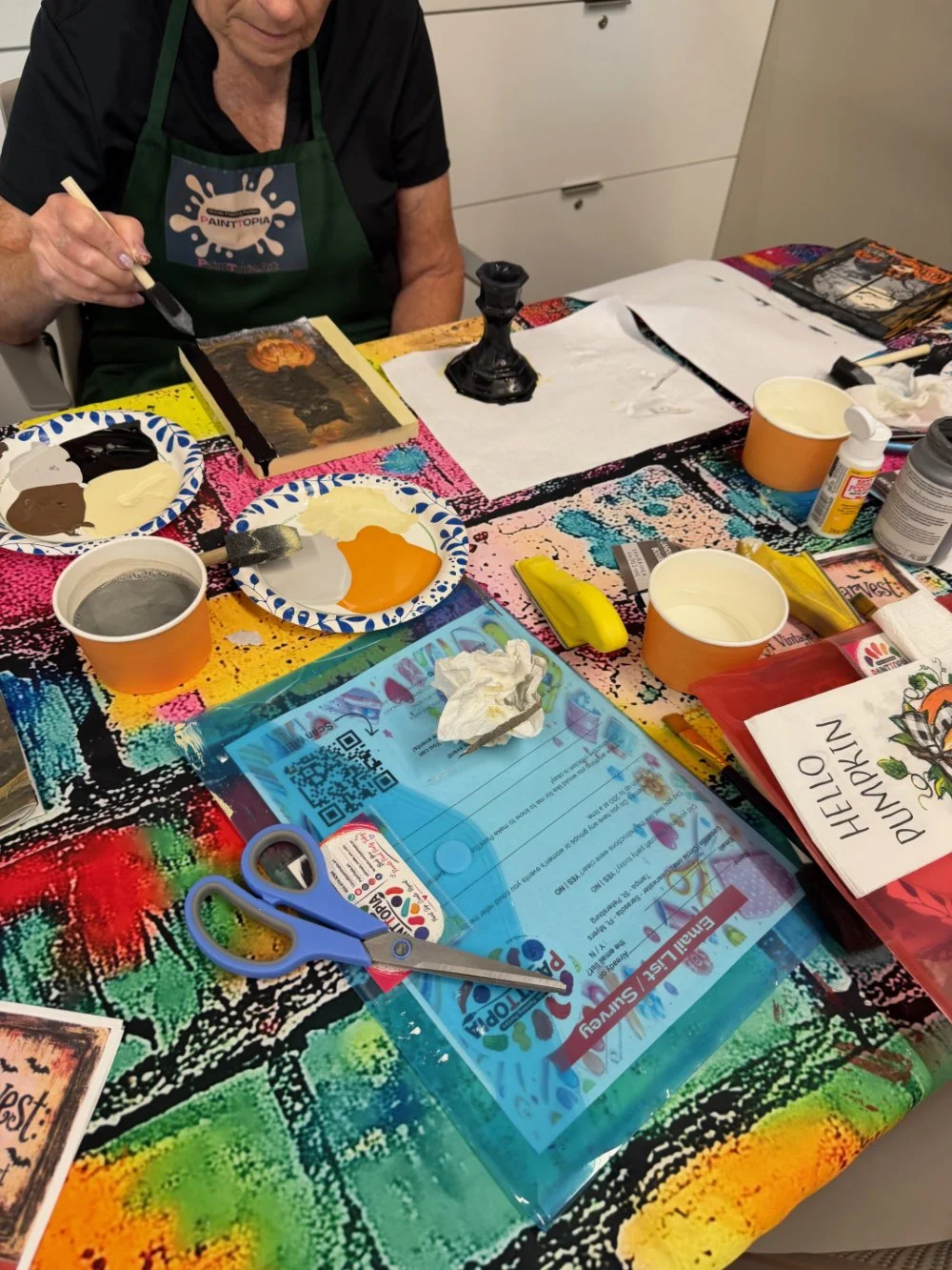 Person painting a small canvas with black and orange paint at a colorful art table. Various painting supplies, paper plates with paint, and magazines on the table.