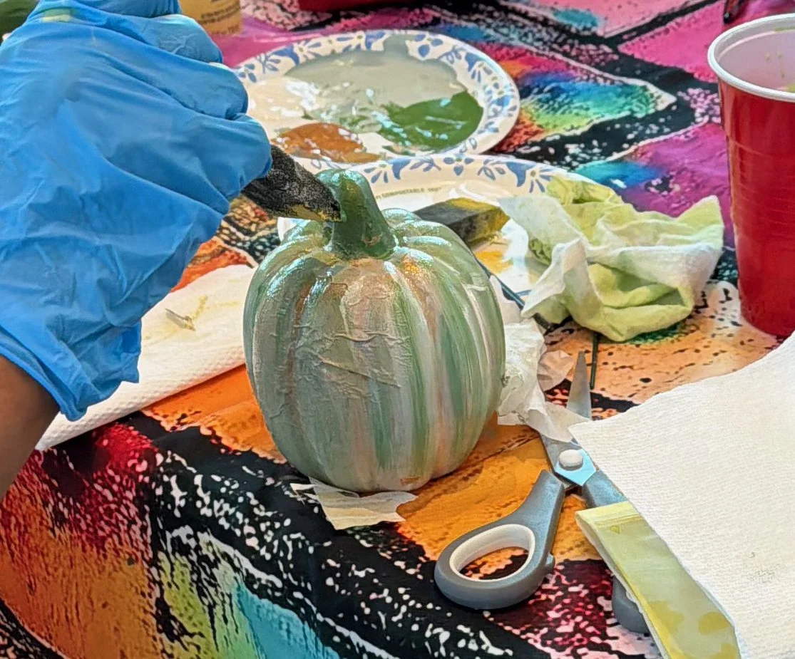 Person painting a small pumpkin with iridescent paint using a black brush, surrounded by painting supplies and paper towels on a colorful tablecloth.