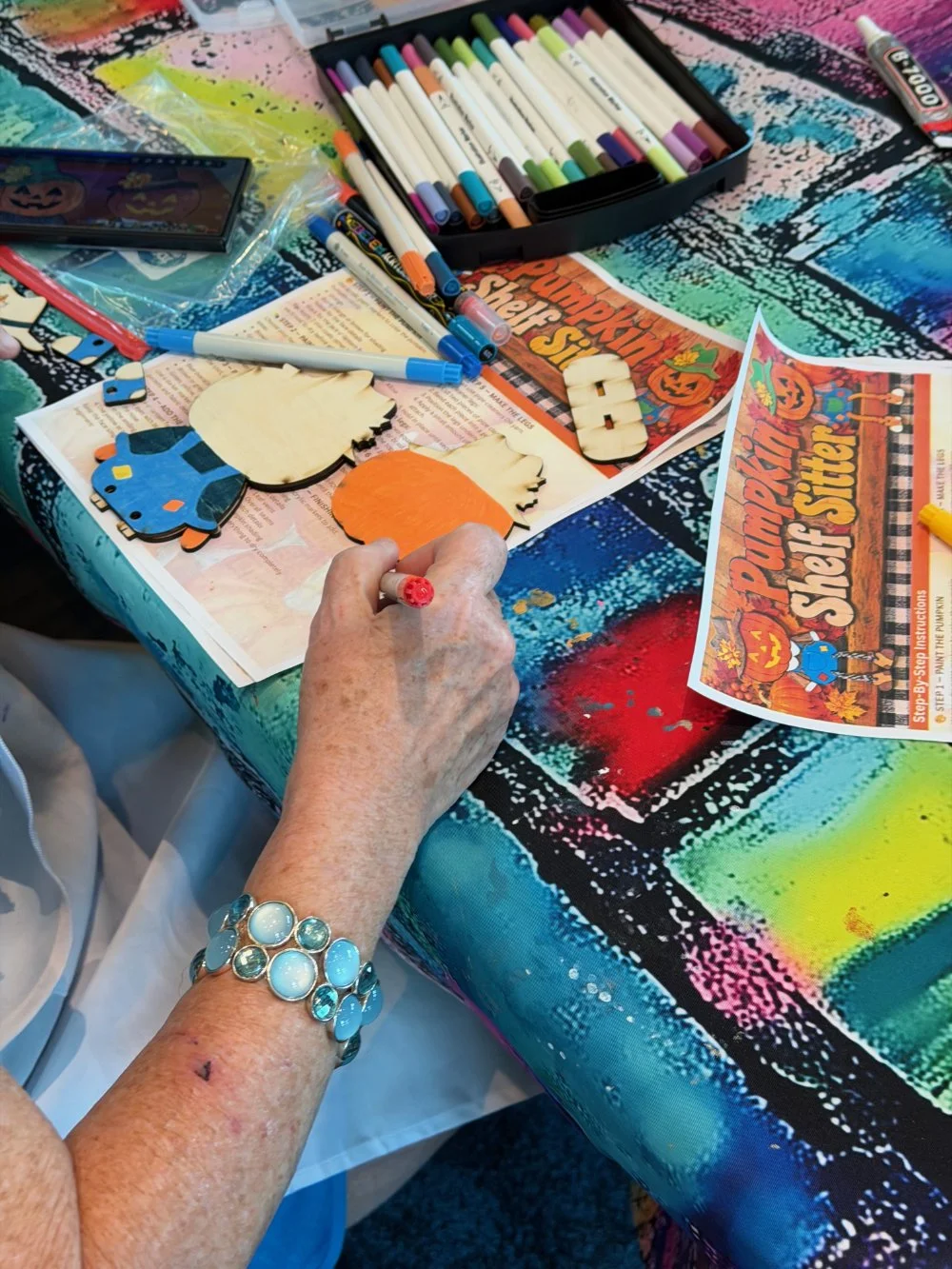 A person with a silver bracelet is creating Halloween-themed wooden cutouts, including a pumpkin, witch, ghost, and a car, at a table with colorful markers, a sheet titled 'Pumpkin Street' with Halloween graphics, and craft supplies for decorating.