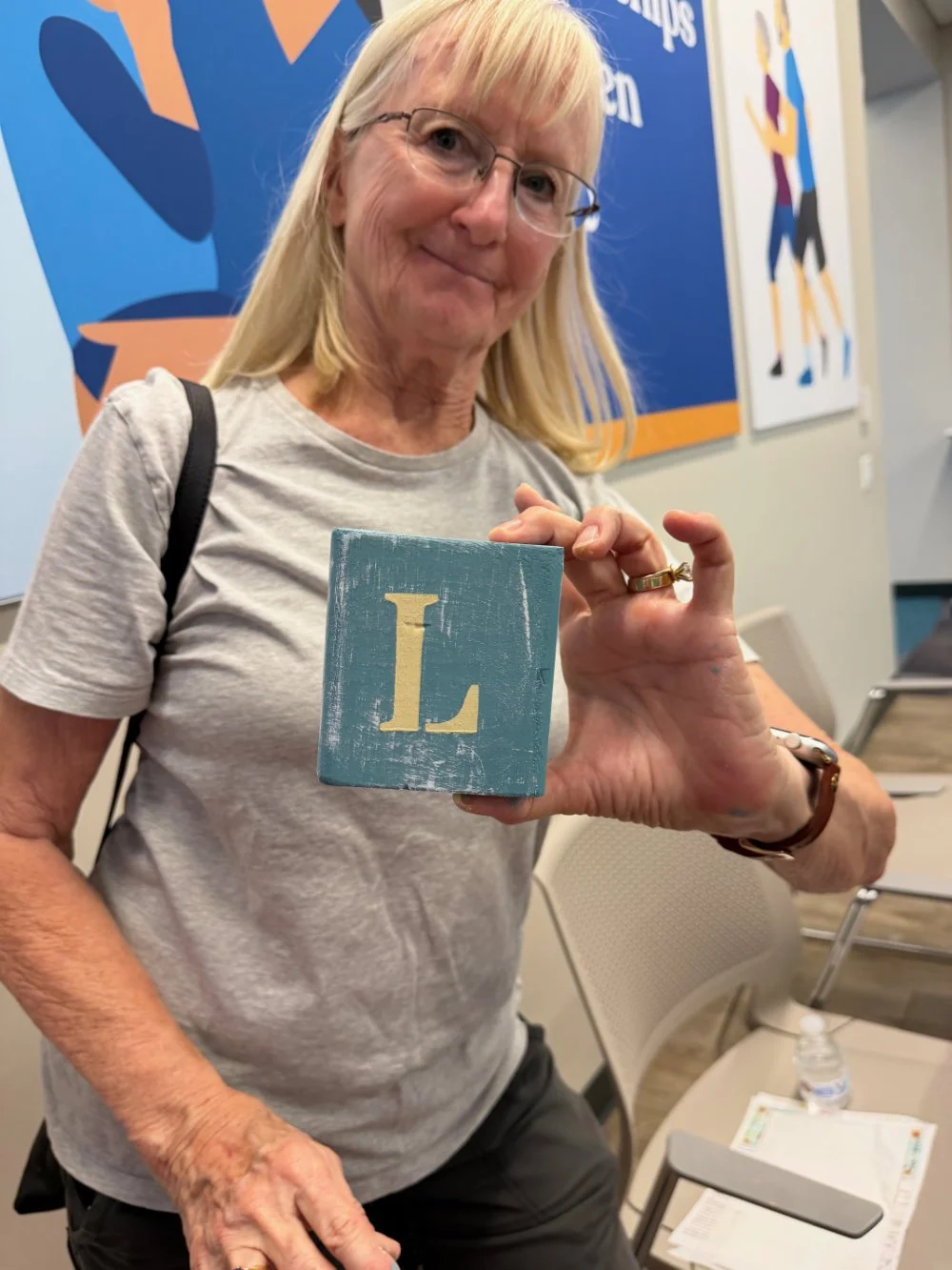 An older woman with blonde hair, glasses, and a Grey t-shirt holds a small blue wooden block with a yellow letter 'L', smiling at the camera, in a room with colorful artwork on the walls.