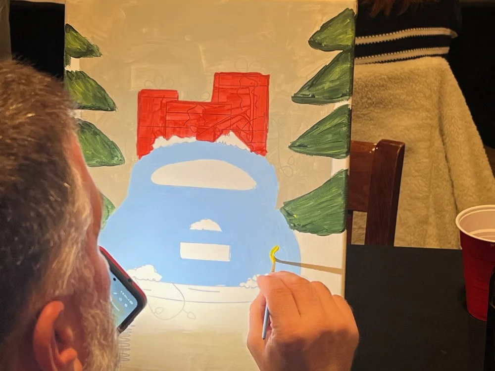 A man painting a snow scene on a canvas, including a snowman with a red brick chimney, green trees, blue snow, and a stick with a yellow end.