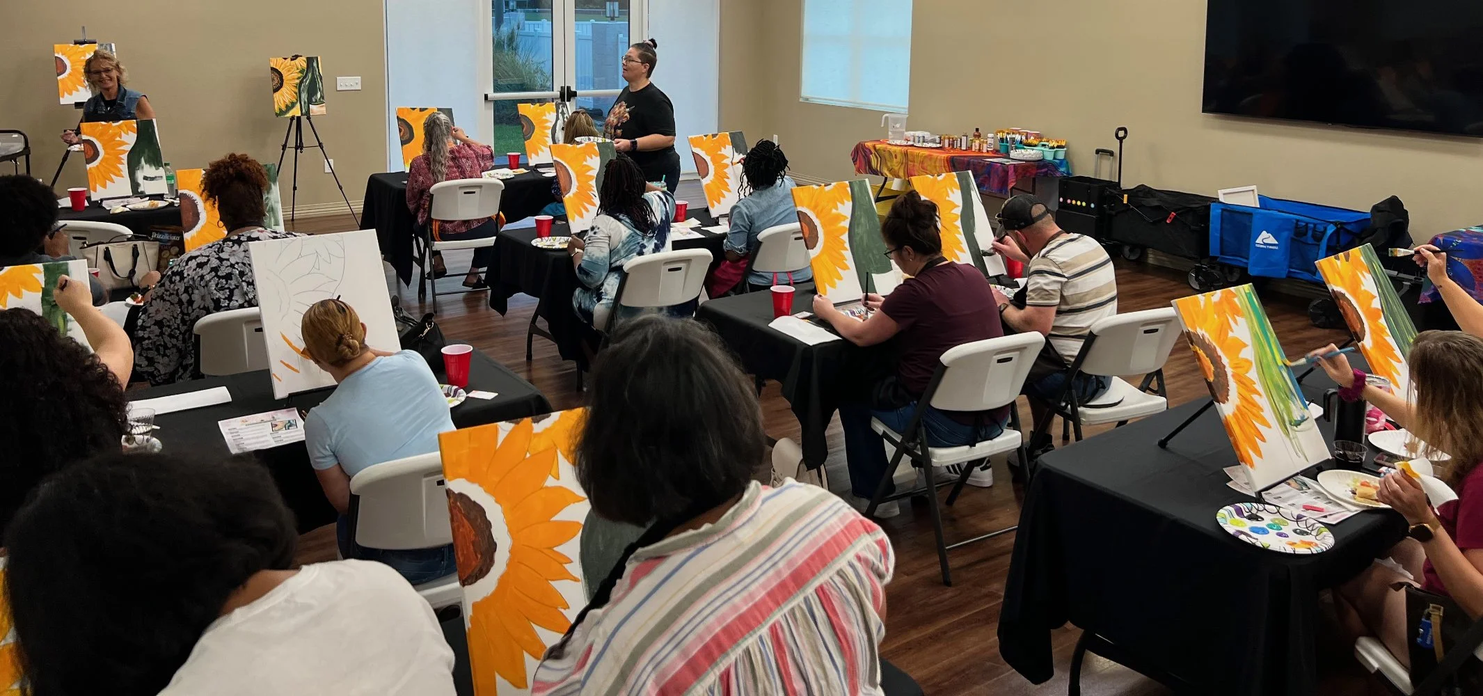 A group of people participating in a painting class, sitting at tables with canvases of sunflower paintings, while an instructor guides them. There are art supplies and refreshments on a table along the wall.