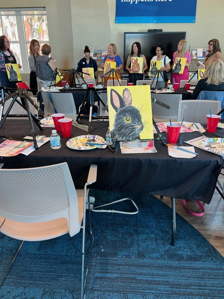 A group of people participating in a painting class, standing behind tables with artist canvases of rabbits and a black cat, set up with paint supplies and red cups.