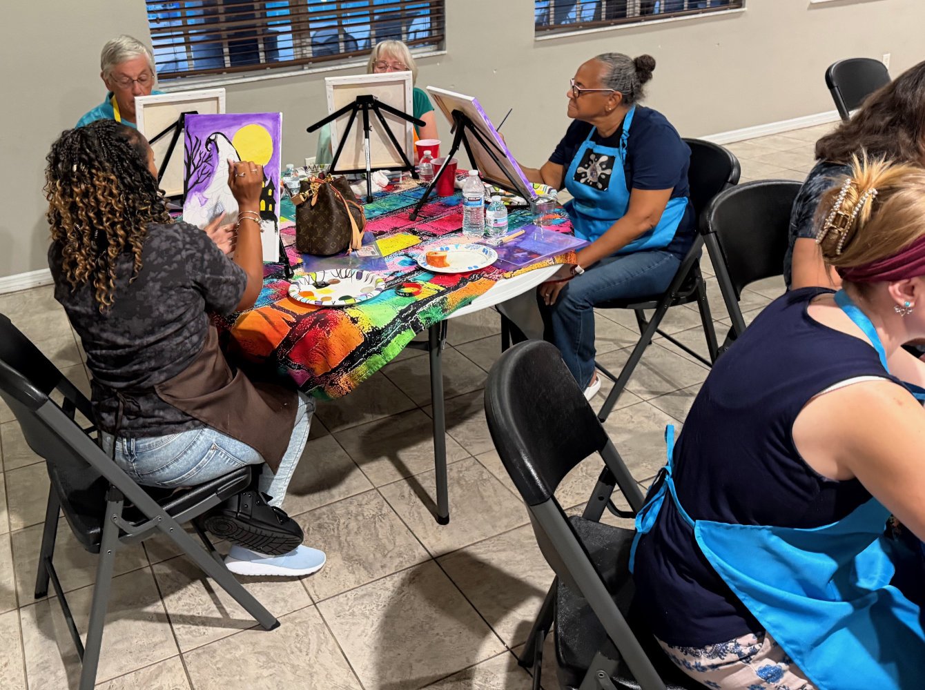 Women participating in a painting class, sitting around a table covered with a colorful cloth, working on canvases with easels.