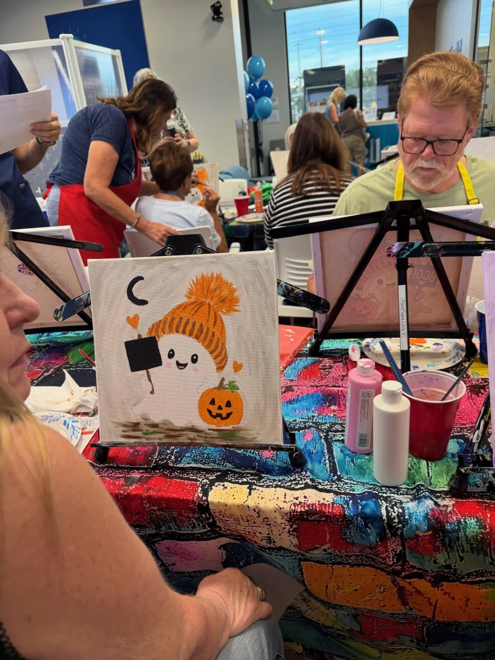 A group of people participating in a painting class at a table covered with a colorful tablecloth. In the foreground, a small canvas depicts a cute ghost wearing a knit hat, holding a black sign, with a pumpkin, hearts, and a crescent moon. The scene
