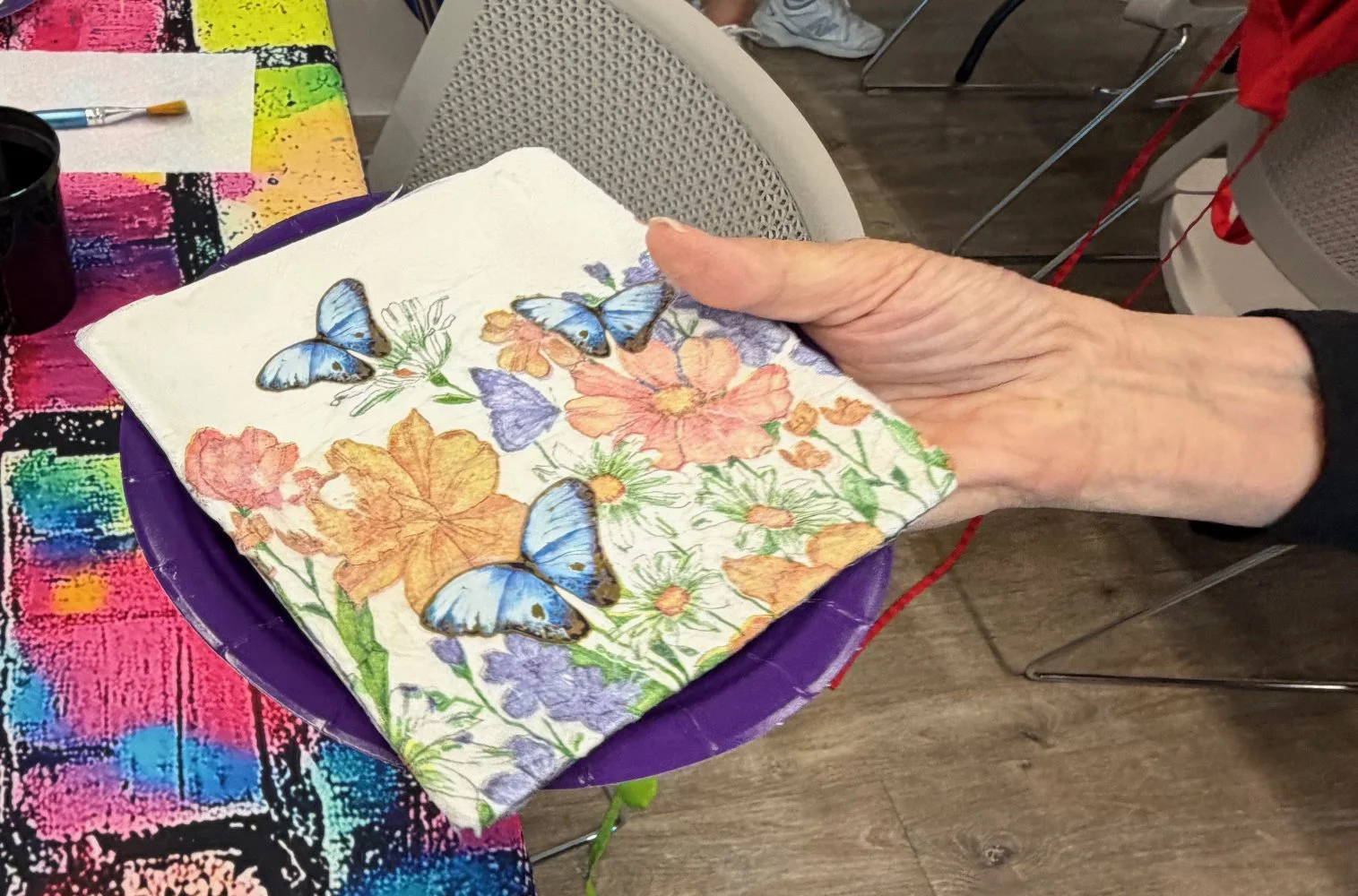 A hand holding a piece of paper with butterfly and floral illustrations, over a purple bowl on a colorful table, with a chair and part of a red jacket visible in the background.