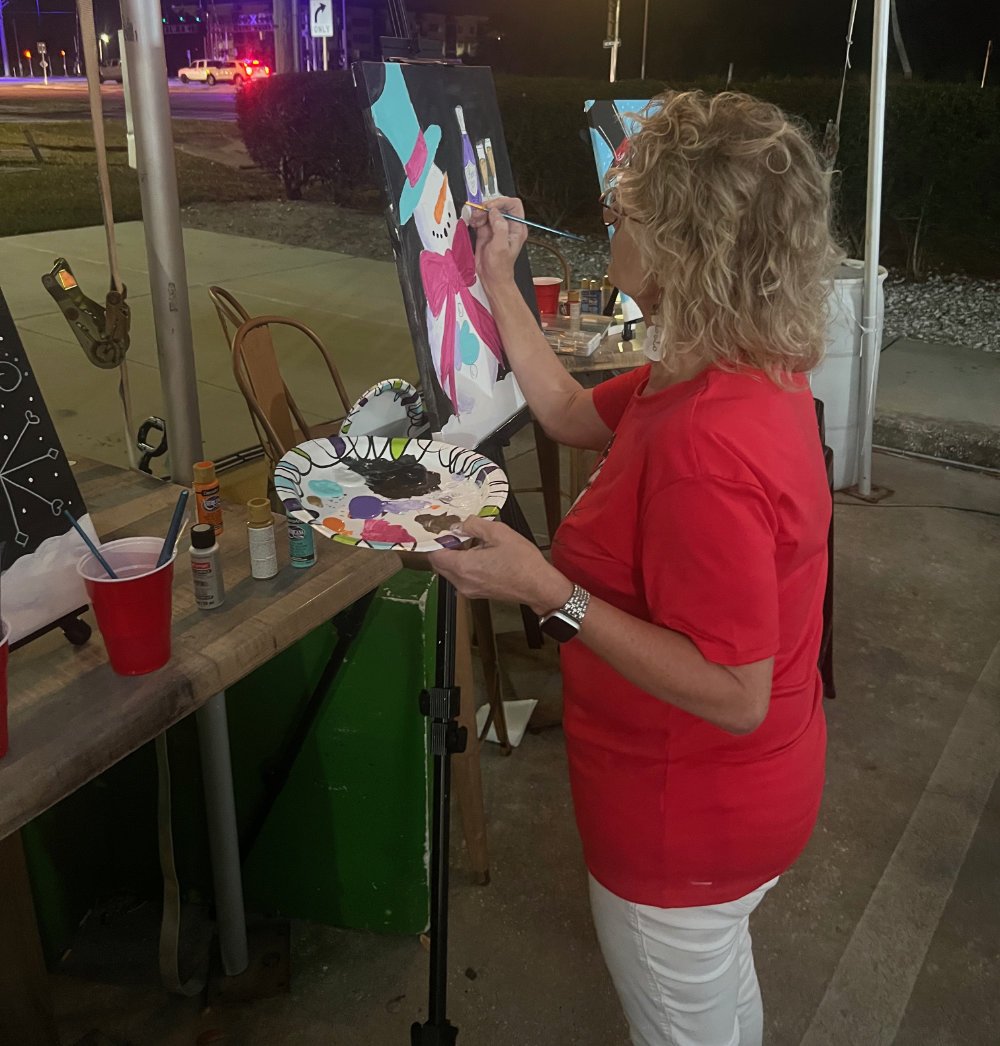 A woman with curly blonde hair painting a colorful abstract piece on an easel at an outdoor art event during nighttime. She is wearing a red shirt and white pants, holding a palette with black paint and using a brush to add details to her artwork.