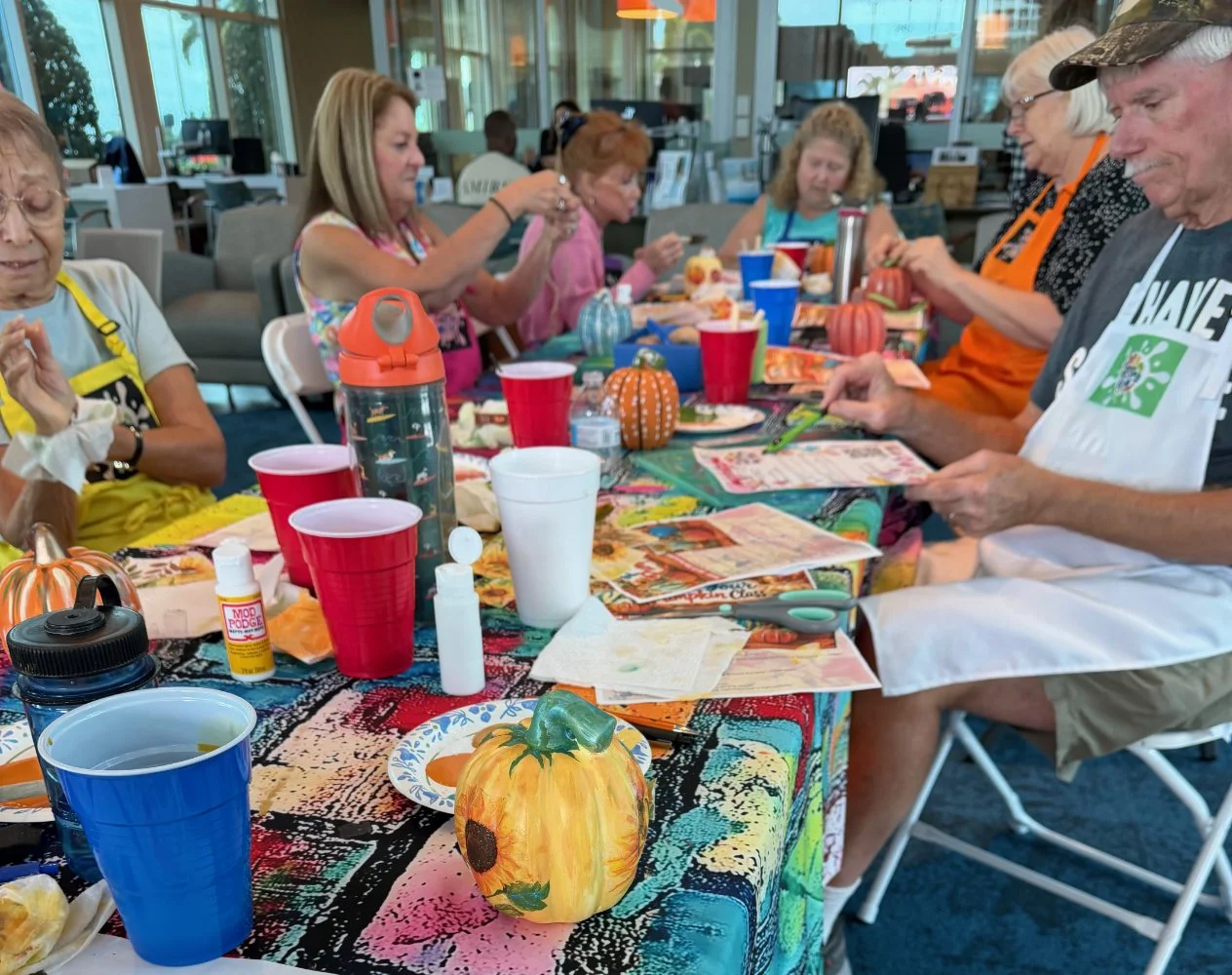 A group of elderly women gathered around a festive table decorated with small pumpkins, eating and reading menus. The table has colorful tablecloths, drinks in red cups, and various craft supplies, suggesting a fall or Halloween-themed gathering at a