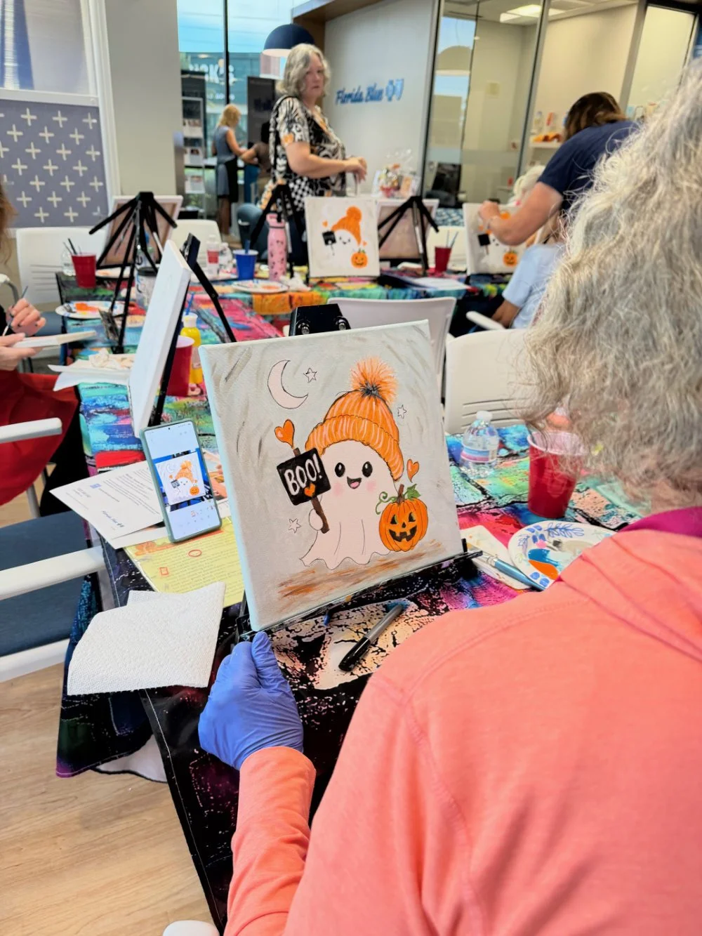 A woman painting a Halloween-themed illustration of a cute ghost with a pumpkin and a pumpkin hat, at an art class, with other people engaged in painting in the background.