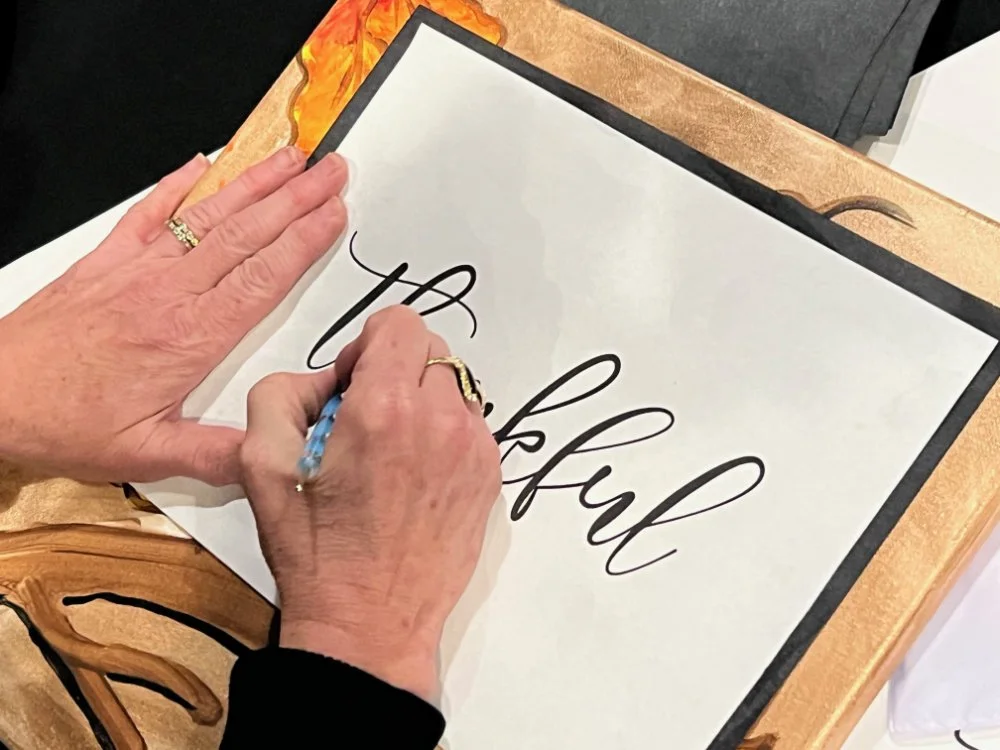 Person signing a framed calligraphy print that reads 'Thankful' with a blue pen. The person is wearing rings and has a gold bracelet.