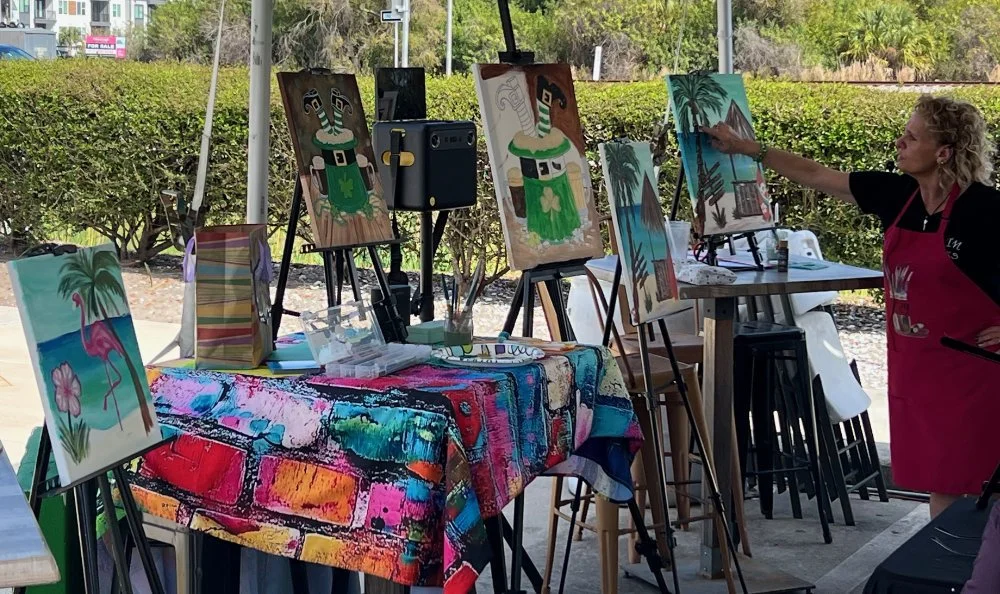 An outdoor painting class with multiple canvas paintings on easels, depicting tropical and beach scenes, with a woman in a red apron painting a canvas and a table with colorful cloth in front.