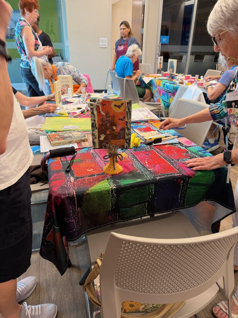 People participating in a craft workshop around a table with colorful tablecloths, creating artwork with various art supplies in a well-lit indoor space.