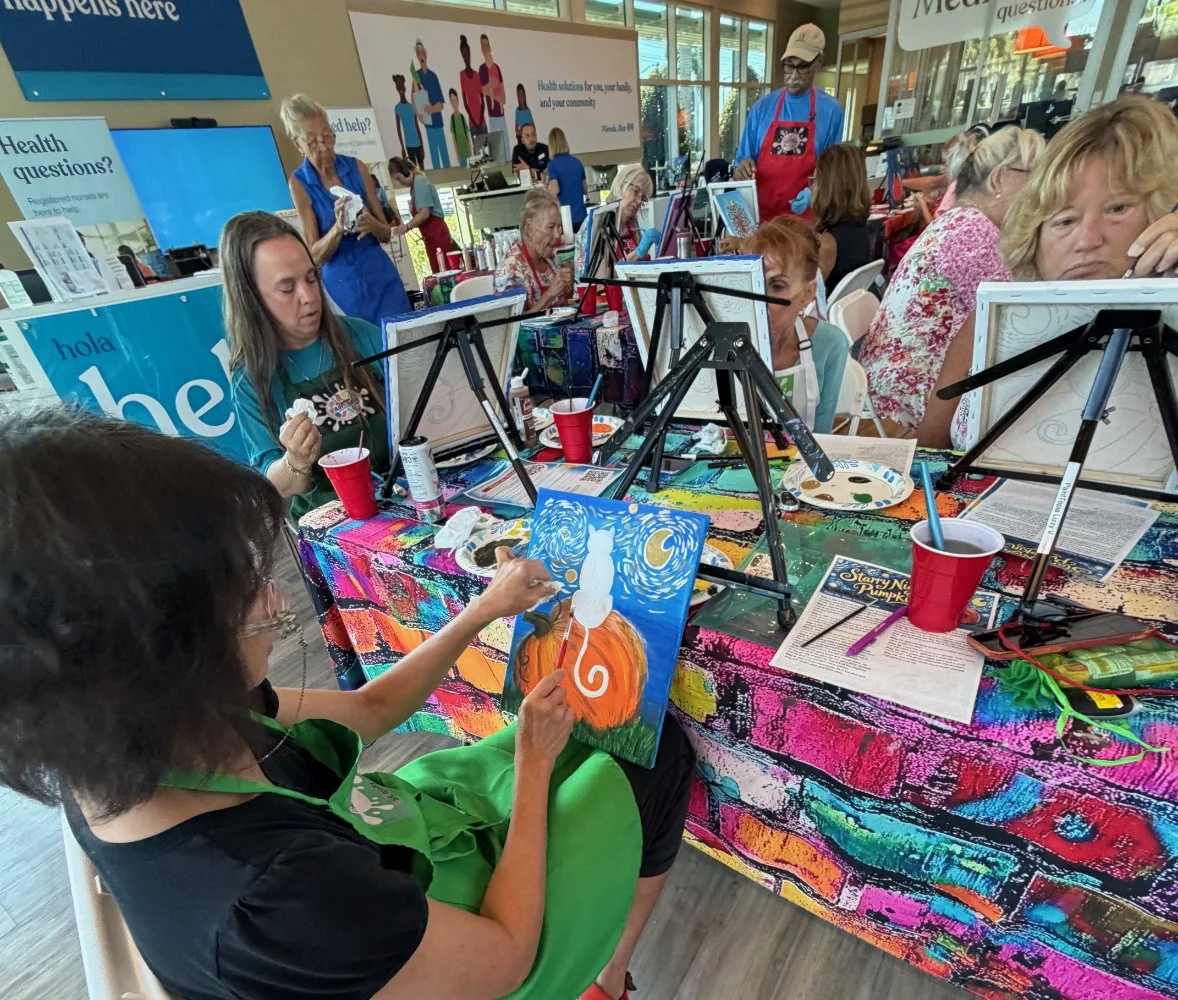 A group of people participating in a painting class, with some painting Halloween-themed artwork featuring a white cat on an orange pumpkin, set up at a colorful table with art supplies in an indoor space.
