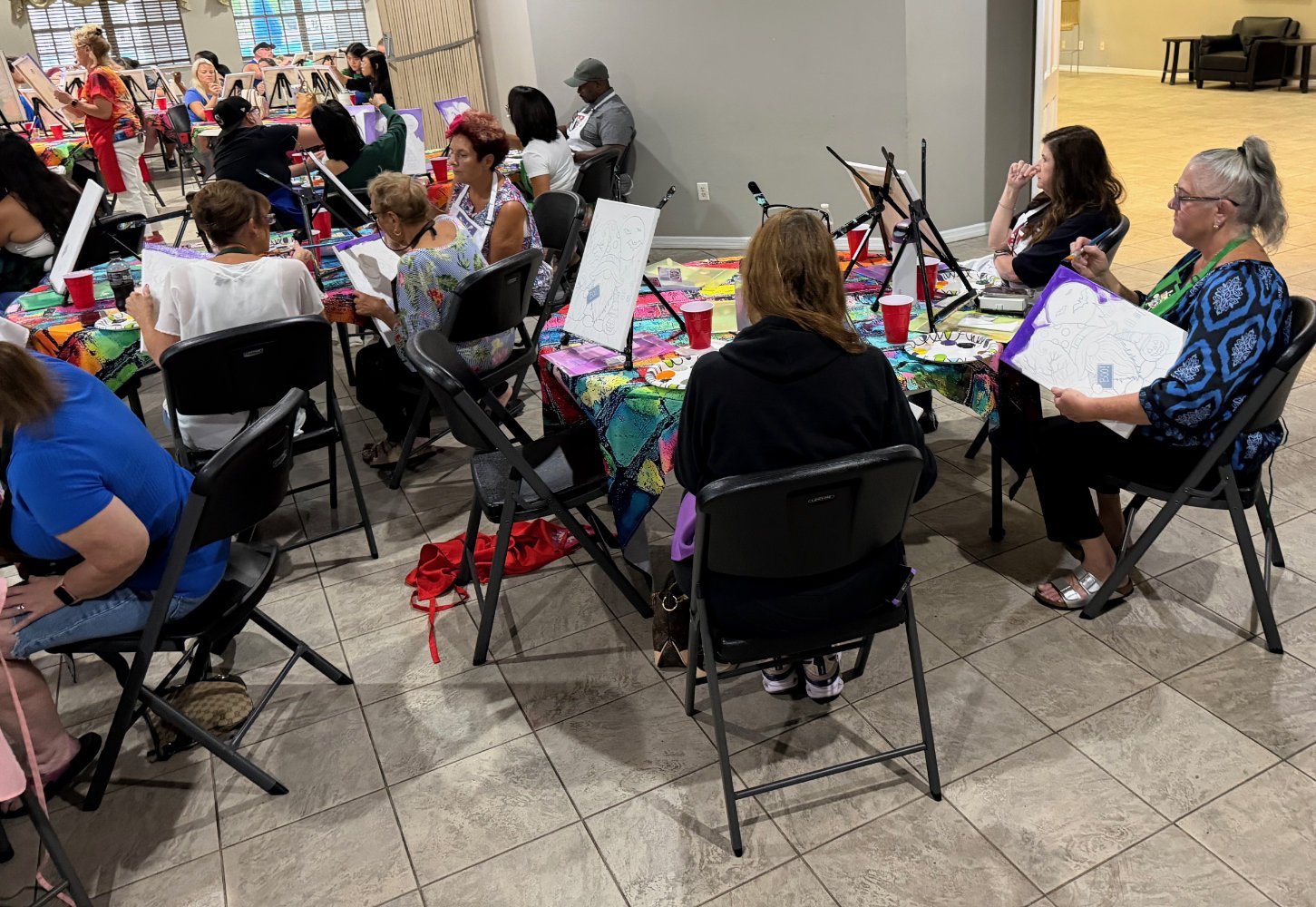 Group of women participating in a painting class, sitting at tables covered with colorful tablecloths, with canvases, easels, and painting supplies, in an indoor community space.