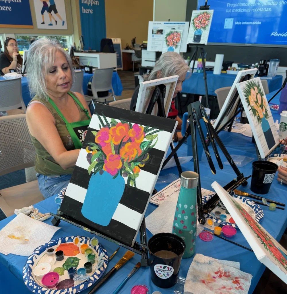 A woman painting a colorful floral still life in a blue vase during a painting class. The table has art supplies, paint palettes, brushes, and drinks. Other participants and easels with paintings are visible in the background.