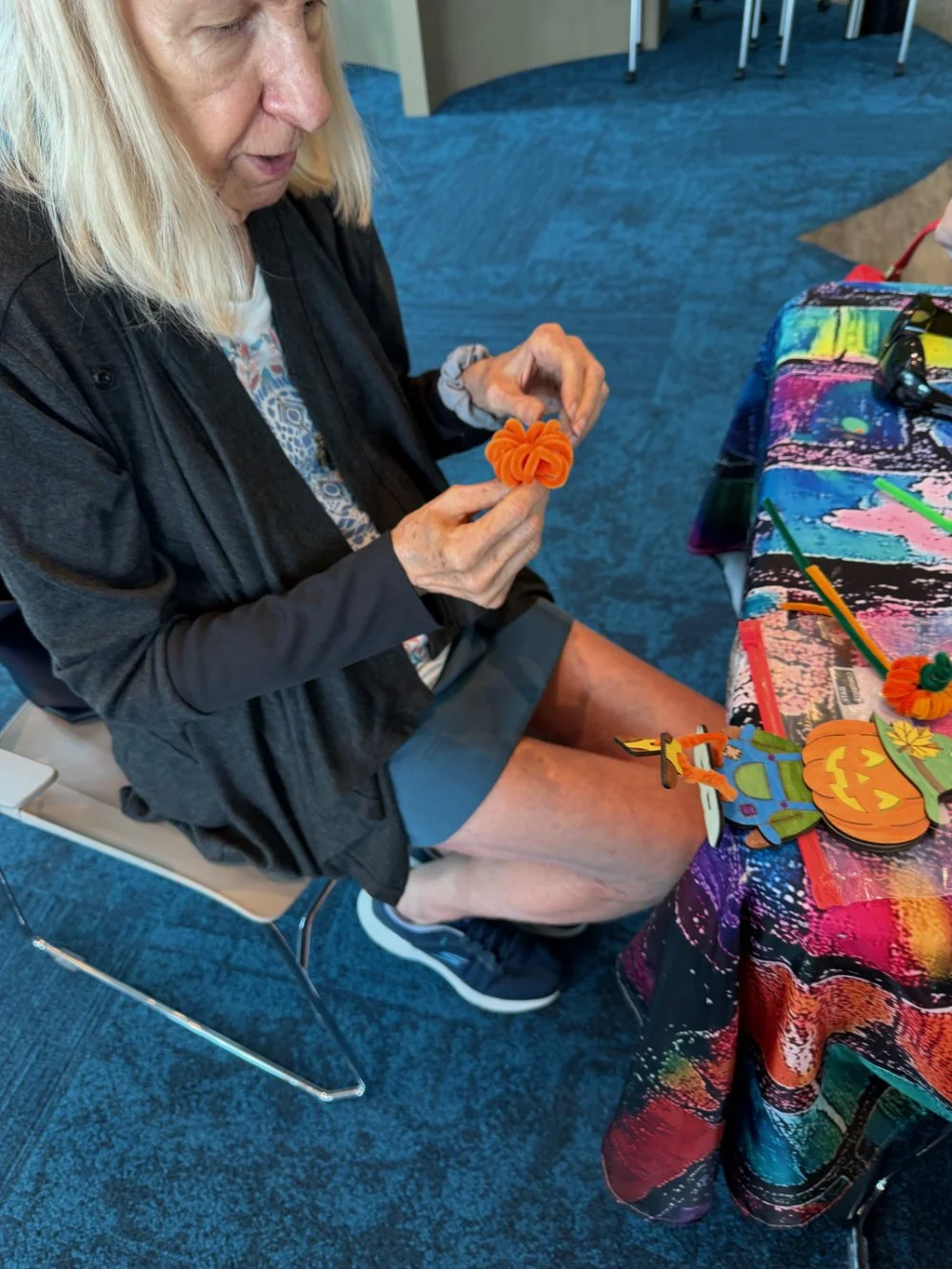 Older woman with blond hair, wearing a black jacket and gray shorts, sitting at a colorful, tie-dye table, holding an orange felt flower craft. Craft supplies and pumpkin decorations are on the table.