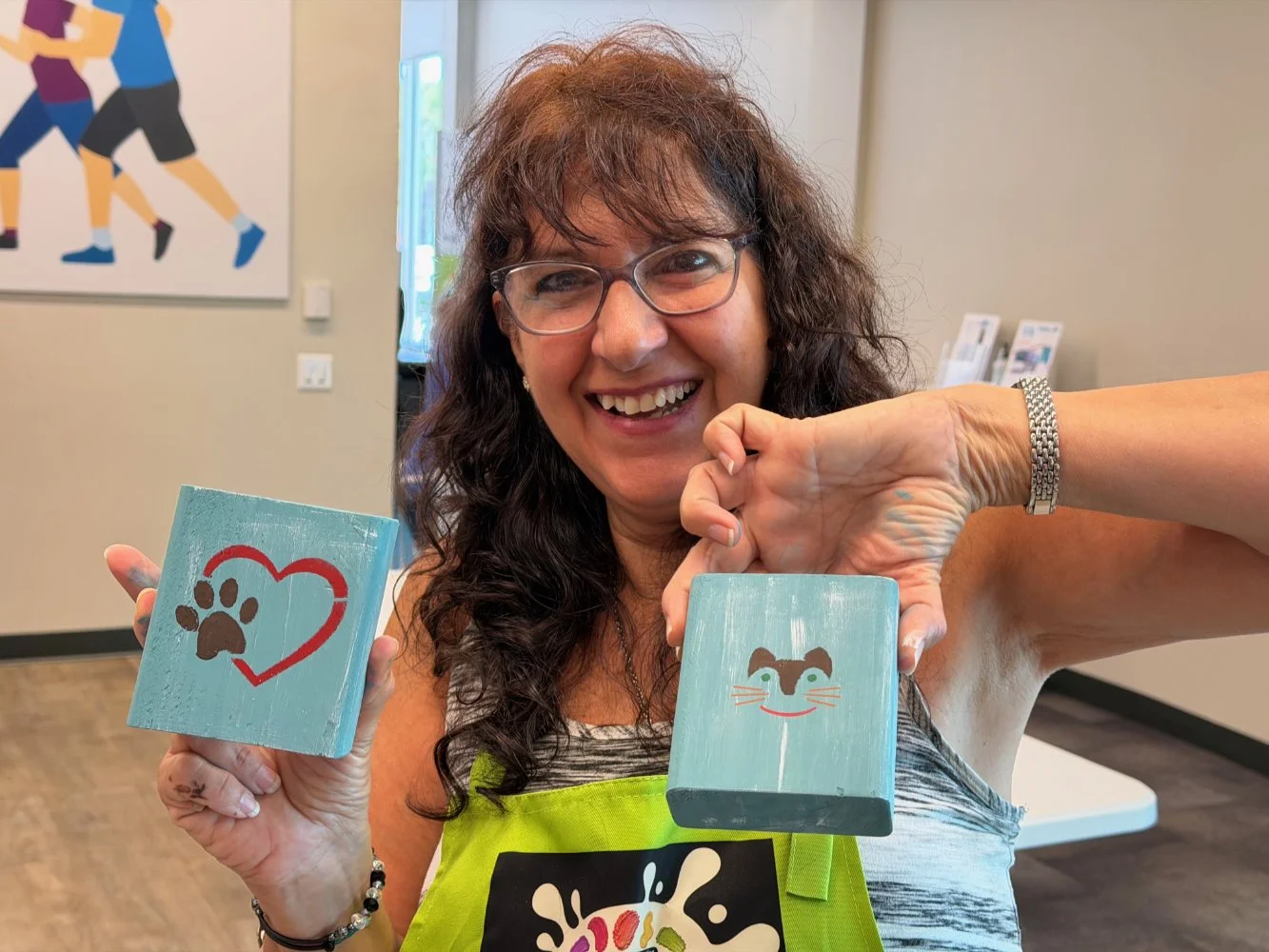 A woman with glasses and curly hair smiling, holding two painted wooden blocks with animal and heart designs inside a room with beige walls and artwork in the background.