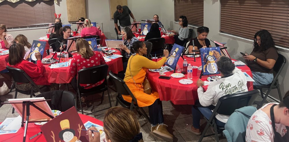 Group of people participating in a paint and sip event, painting snowmen on canvases at decorated tables.