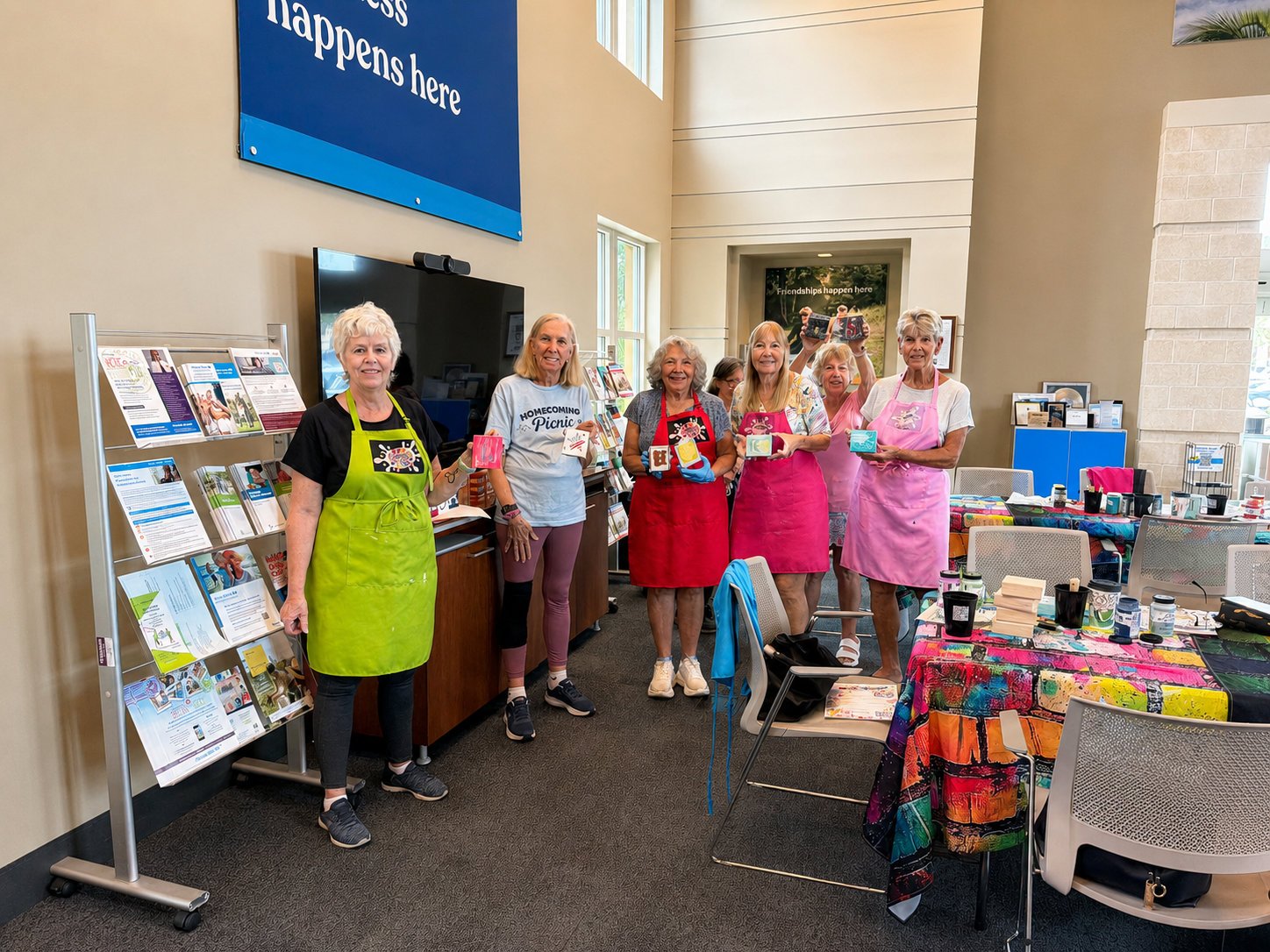 Group of six women, some wearing pink aprons, standing inside a community center or event space, holding small boxes, with tables of art supplies and informational materials around them.
