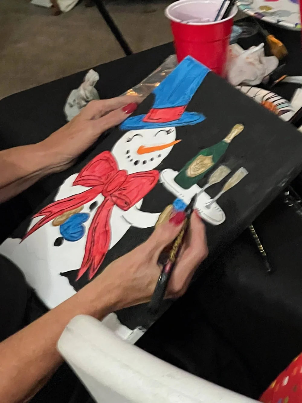 Person painting a snowman on a black canvas, wearing a blue hat with red band, a red scarf, and using a paintbrush. The snowman has a carrot nose, black eyes, and mouth, with a bottle of champagne and glasses nearby.