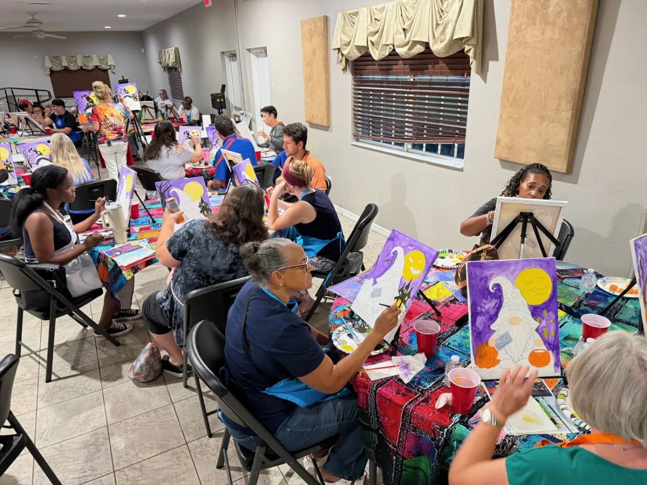 People participating in a group painting class, sitting around tables with canvases of witches, moons, and pumpkins in a well-lit room with beige walls and windows.