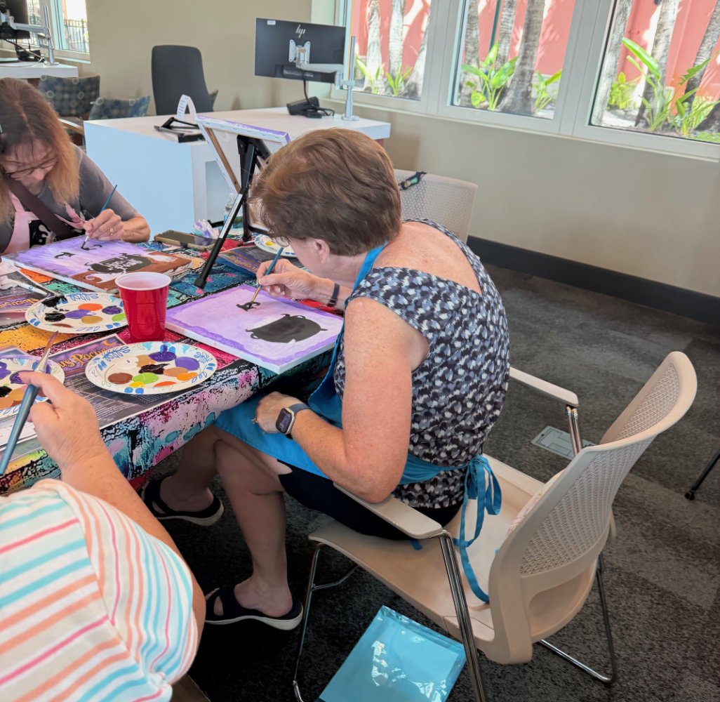 People painting at a table with art supplies in a bright room with large windows.